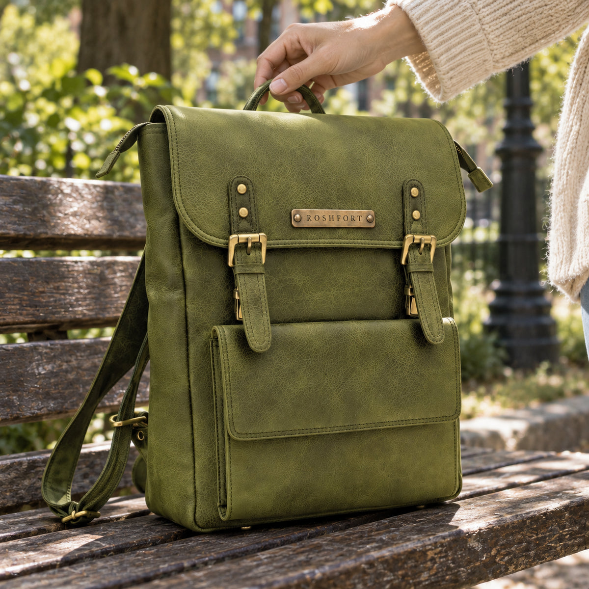 The olive Italic Royal backpack on a park bench, being picked up by its top handle