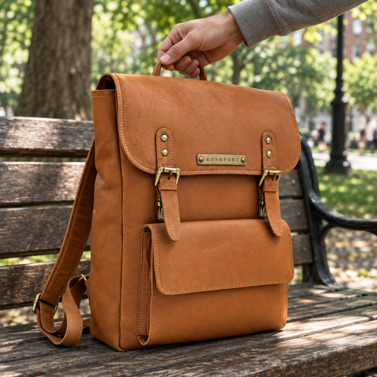 The tan Italic Royal backpack on a park bench, being picked up by its top handle